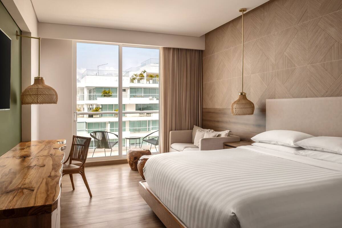 Een moderne hotelkamer met een groot bed, een houten bureau en een balkon met uitzicht op andere gebouwen.