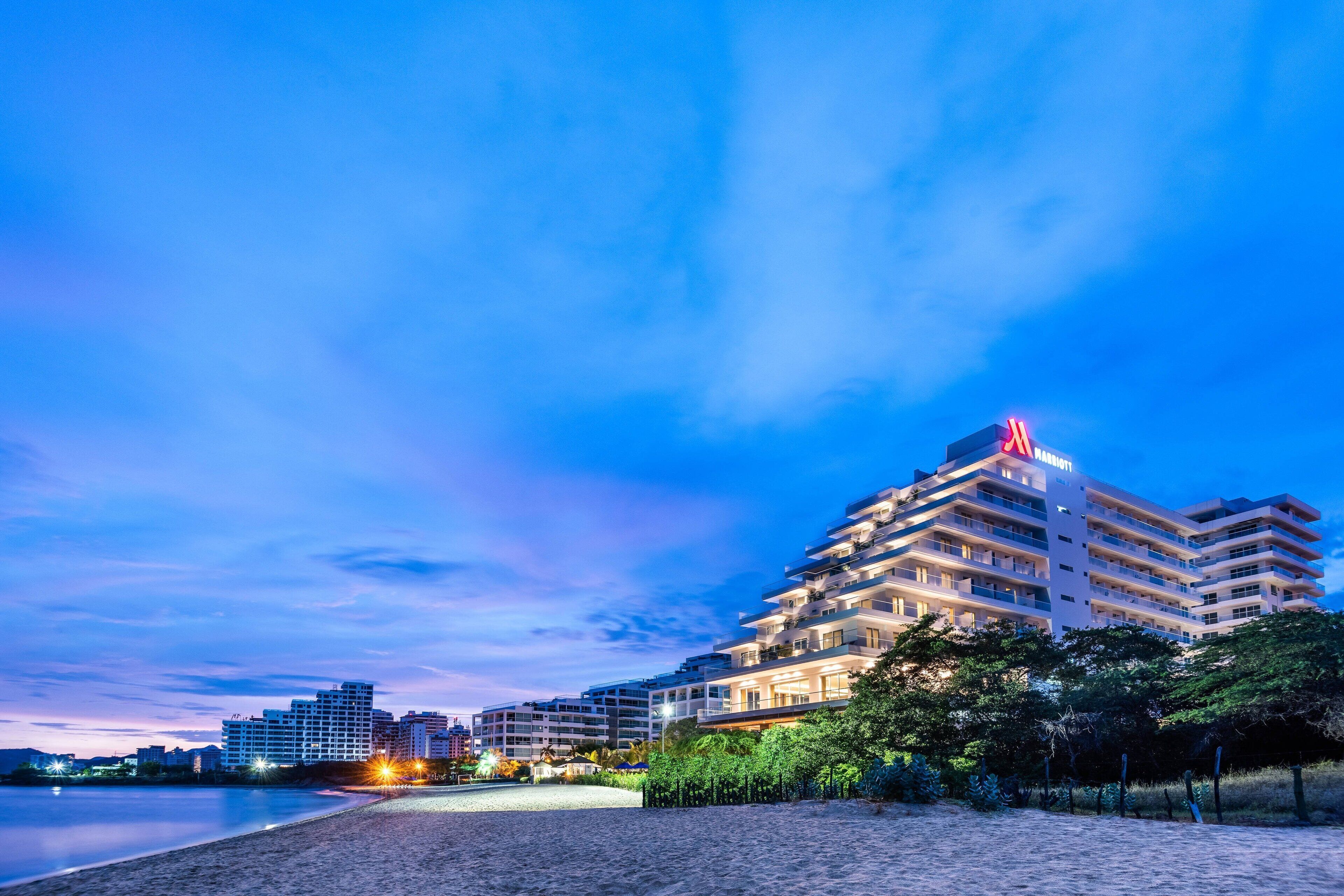 A multi-story hotel with a prominent sign, located near a beach with a calm sea and a clear sky.