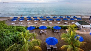 A beach with rows of white lounge chairs and blue umbrellas, palm trees, and a view of the sea.