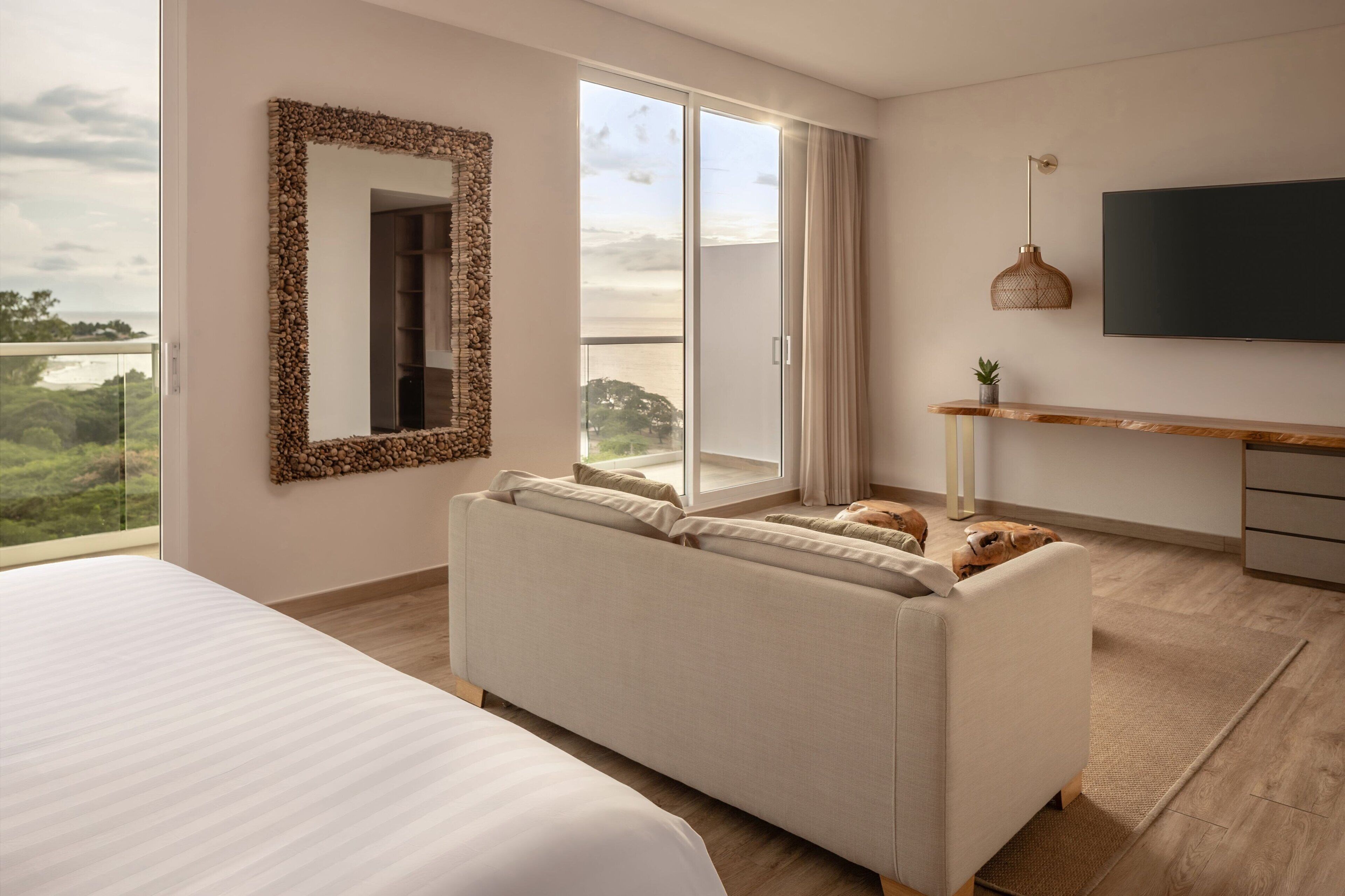 A modern hotel room with a large bed, a sofa, a TV, and a balcony with a view of the outdoors.