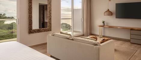 A modern hotel room with a large bed, a sofa, a TV, and a balcony with a view of the outdoors.