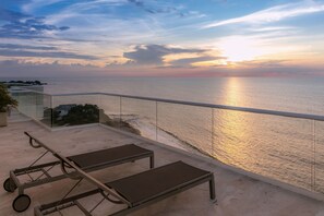 A balcony with a view of the sea at sunset, a glass railing, and two sun loungers.