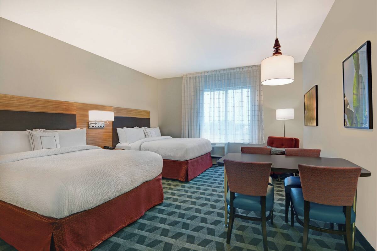 Quarto de hotel com duas camas, uma mesa de jantar com cadeiras, uma janela grande com cortinas e um quadro na parede.