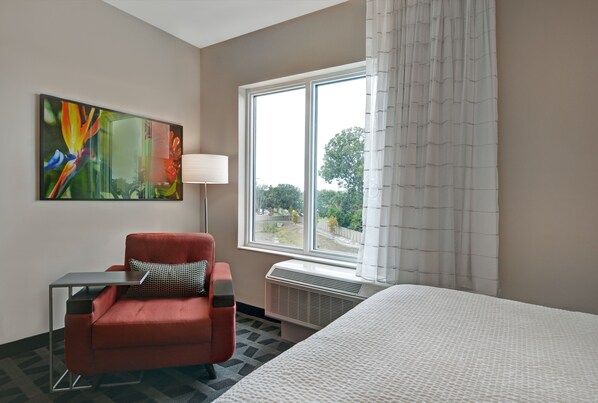 A hotel room with a bed, a red armchair, a small table, a lamp, and a large window with curtains.