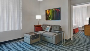 Habitación de hotel con sofá gris, una mesita, lámpara, un cuadro y una cama con colcha roja.