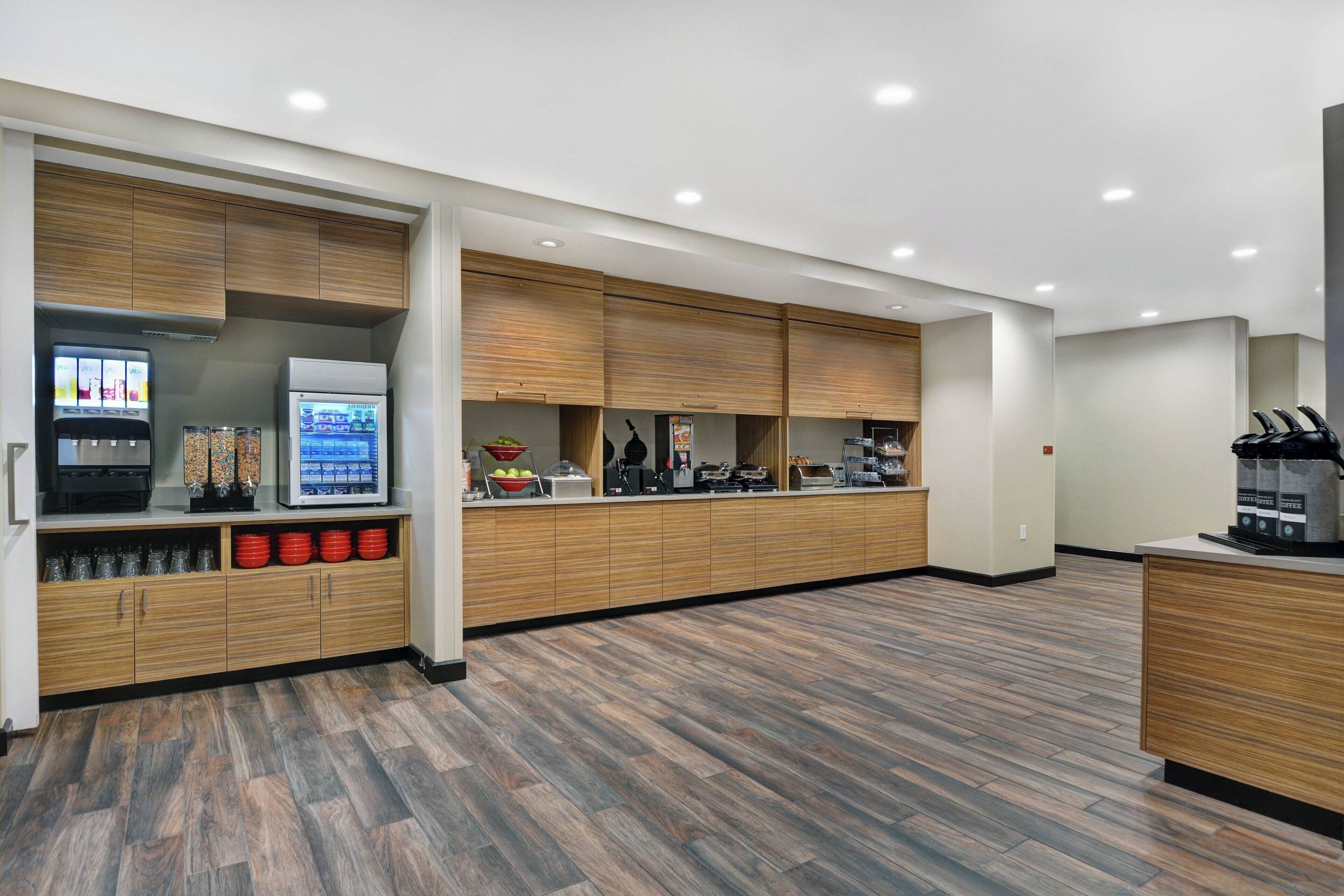 A modern kitchen with wooden cabinets, a refrigerator, a coffee machine, and a beverage dispenser.
