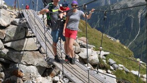 A wooden suspension bridge over a rocky stream with a mountainous backdrop.
