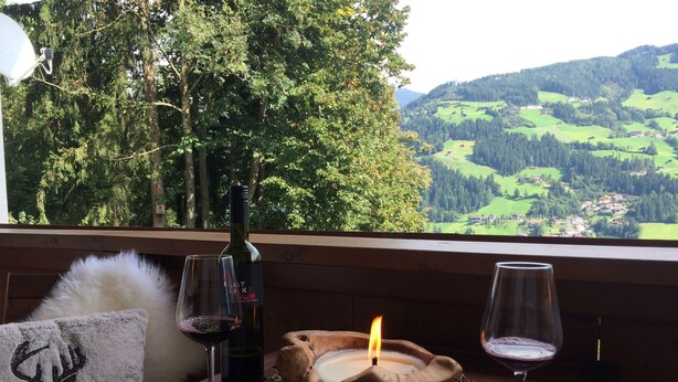 A wooden table with a candle, two wine glasses, and a bottle of wine on a balcony overlooking a scenic landscape.