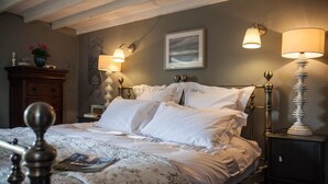 Een netjes opgemaakt bed met witte lakens, twee bedlampjes, een gekaderd kunstwerk aan de muur, en een houten nachtkastje met een lamp.