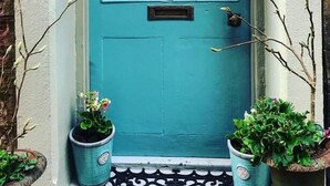 Een blauwgroene deur met een bloemenkrans, twee potplanten en een lantaarn aan de muur.