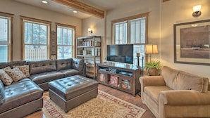A living room with a sectional sofa, ottoman, and a TV stand with shelves and a flat-screen TV. - Updated Townhome w/ Hot Tub - Walk to Downtown! (Red Lodge)