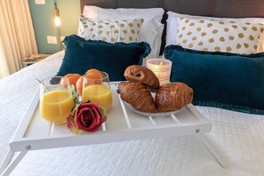 Un vassoio colazione ordinato con croissant, succo d'arancia e una rosa su un letto con cuscini color acquamarina e una testiera bianca.