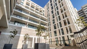 A modern multi-story building with balconies, large windows, and a paved courtyard with planters. - Beautiful 1 bedroom in great central area (Jerusalem)