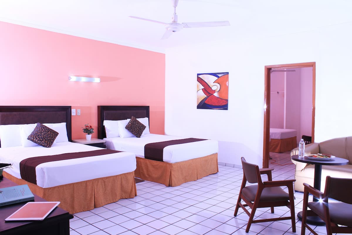 Habitación de hotel con dos camas, un escritorio con una computadora portátil, una silla, una mesa con una botella y un ventilador de techo.