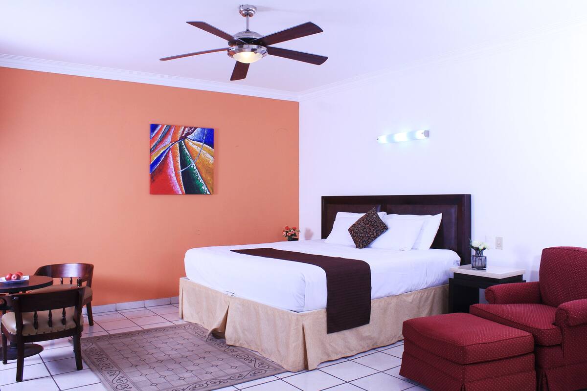 Una habitación de hotel con cama, ventilador de techo, un cuadro en la pared, una mesa de comedor con sillas y un sillón rojo.