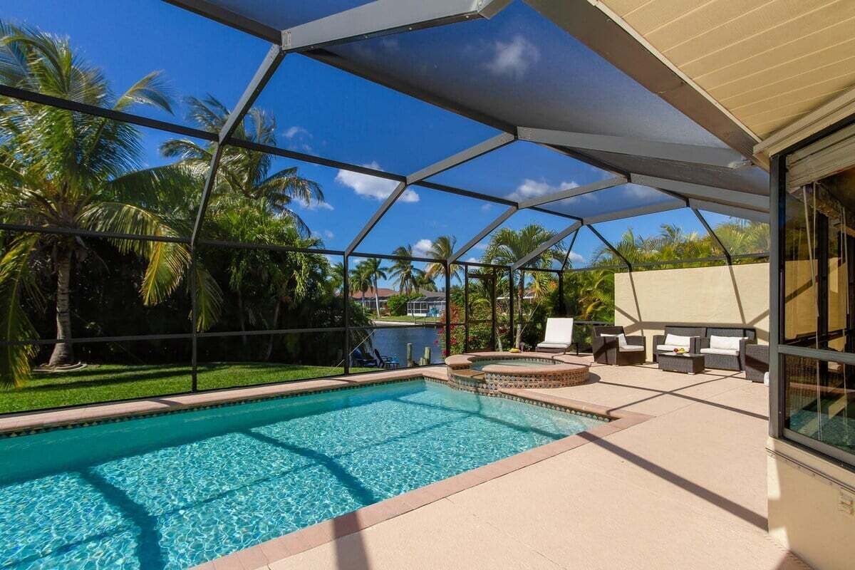 A swimming pool with a covered patio area, lounge chairs, and a view of palm trees.