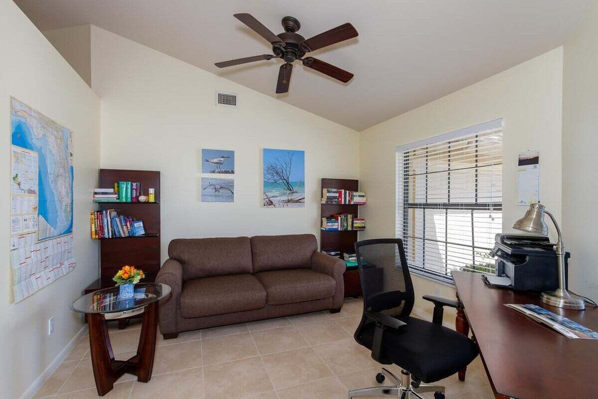 A living room with a brown sofa, a coffee table, a bookshelf, a desk with a computer, and a ceiling fan.