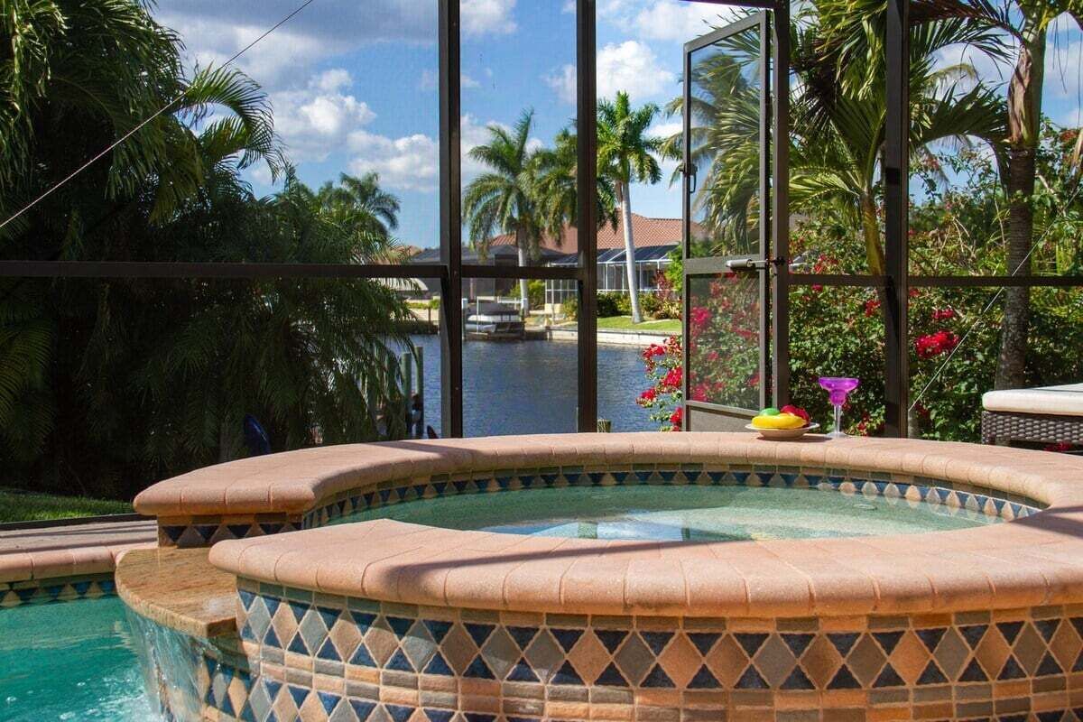 Un jacuzzi con borde de azulejos, rodeado de abundante vegetación y vistas a un cuerpo de agua y un edificio al fondo.