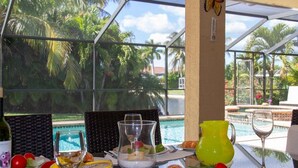 A dining table with a glass pitcher, a wine glass, and a bottle of wine, set against a backdrop of a pool and palm trees. - Ellen`s House-pool and Jacuzzi, Gulf access (Cape Coral)
