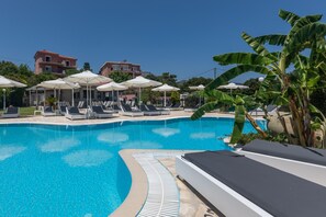 Ein Swimmingpool mit Liegestühlen und Sonnenschirmen, umgeben von Gebäuden und einer Bananenpflanze.