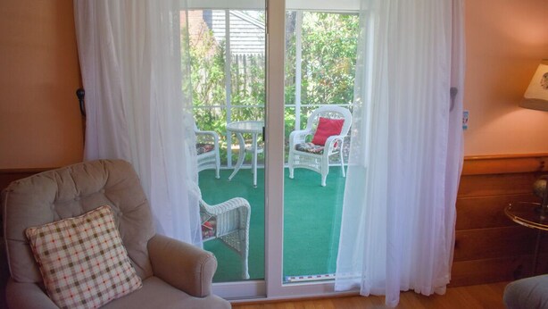 Une pièce dotée d’une porte-fenêtre coulissante donnant sur un patio extérieur aménagé avec des meubles en osier blanc et un tapis vert.