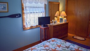 Une chambre à coucher avec un lit, un téléviseur posé sur un meuble, une commode, un miroir et une fenêtre avec des stores.