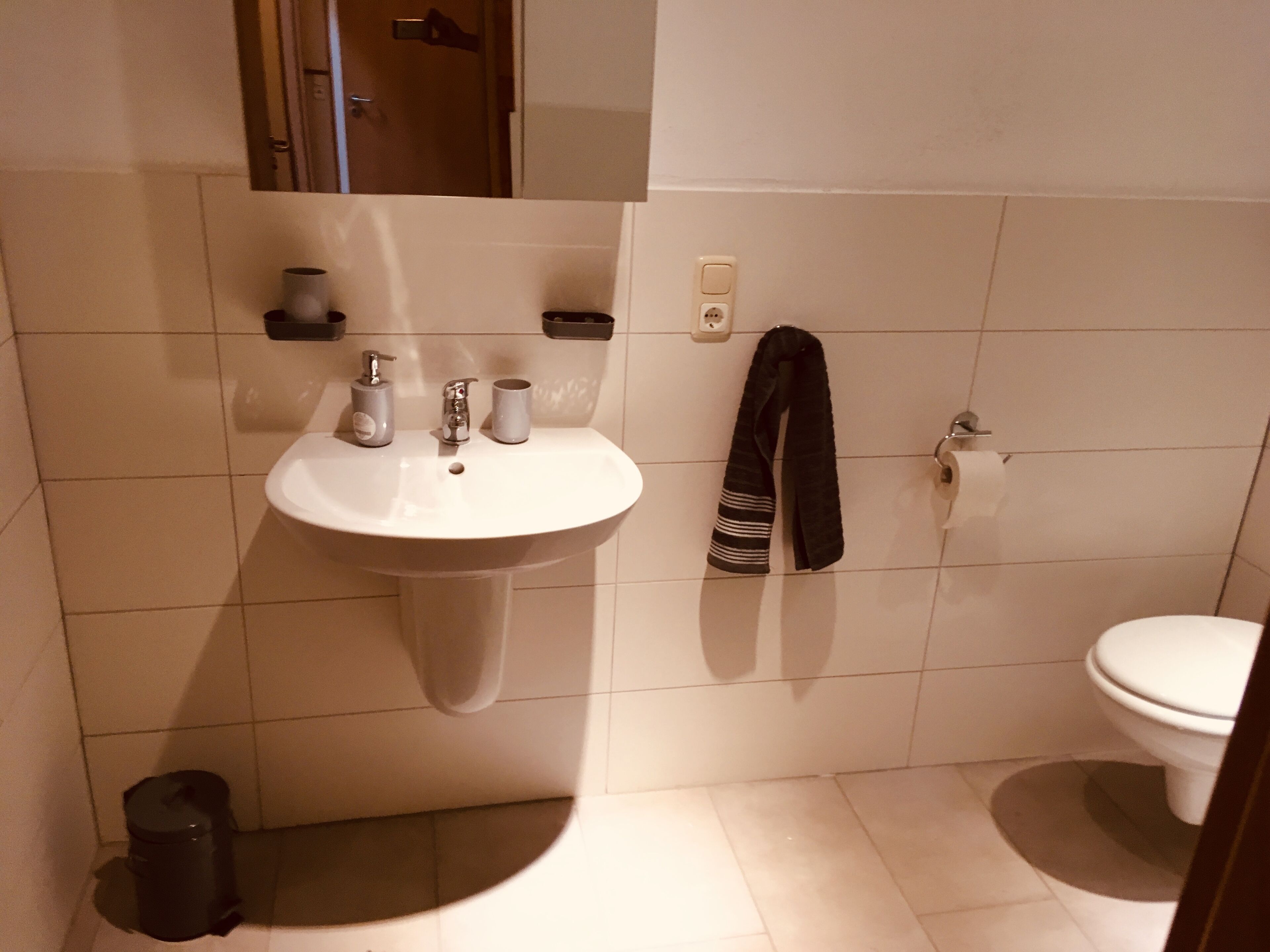 Ett badrum med ett vitt handfat, två kranar, en handduk, en toalett och en papperskorg.
