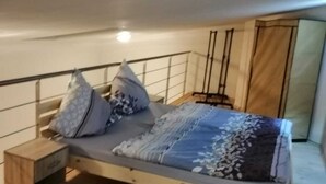 Yhden hengen sänky puukehikolla, yöpöytä ja vaatekaappi huoneessa, jossa on puulattia ja valkoinen katto.