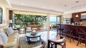 A spacious living area with a sofa, coffee table, and chairs, a kitchen with a bar, and large sliding glass doors leading to a balcony with a view of palm trees and the ocean. - Aloha Villas 3, Oceanfront Townhouse, Steps to The (Kihei)