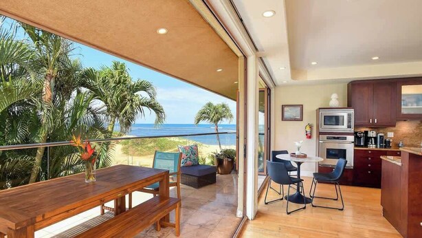 A modern kitchen with a large island, a dining area with a wooden table and benches, and a view of the ocean through large sliding glass doors. - Aloha Villas 3, Oceanfront Townhouse, Steps to The (Kihei)