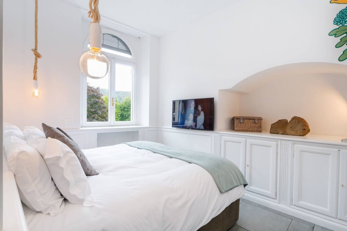 A bedroom with a bed, a window, a television, and a shelf with decorative items.