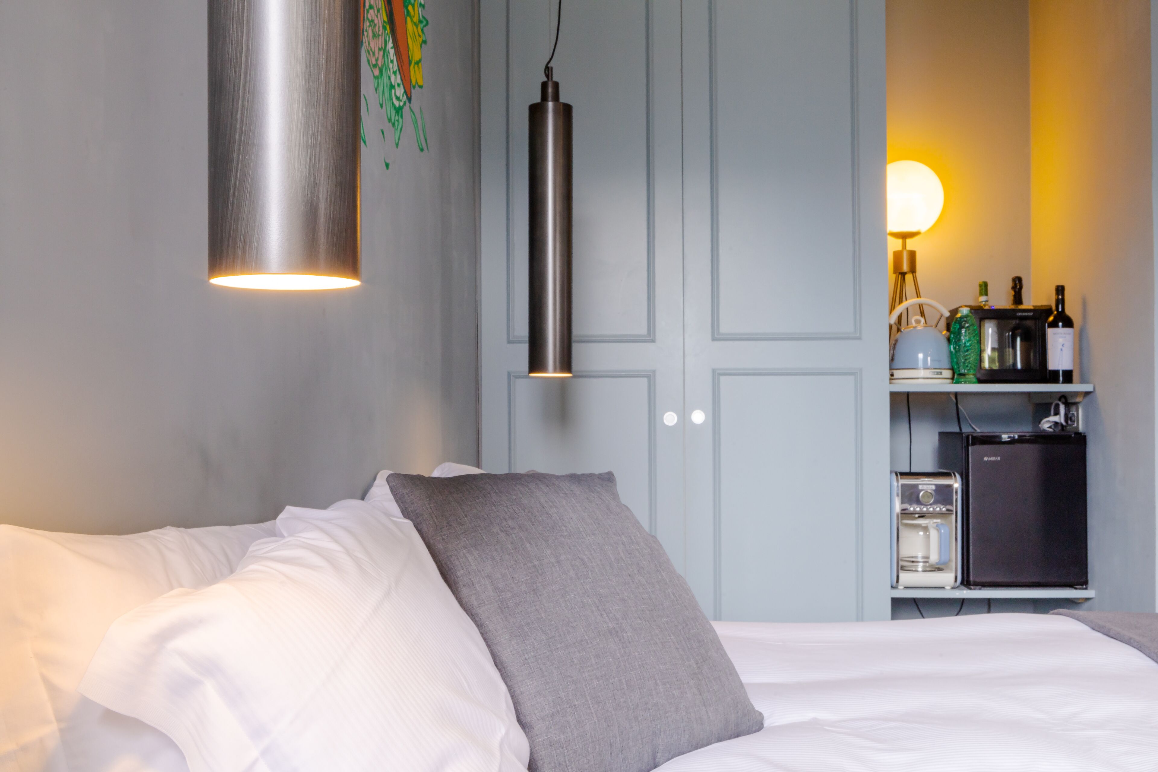 A hotel room with a bed, a grey pillow, a hanging pendant light, a minibar, and a wall-mounted shelf with bottles.