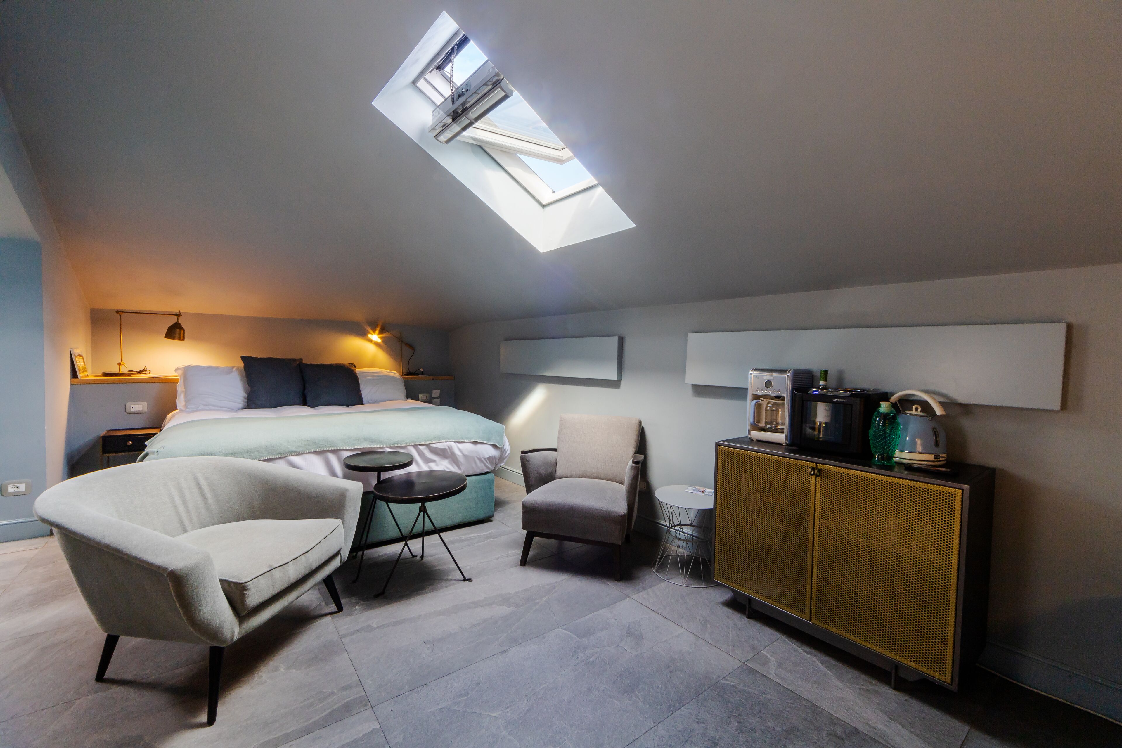 A modern bedroom with a skylight, a bed with pillows, a chair, a small table, and a cabinet with items on top.