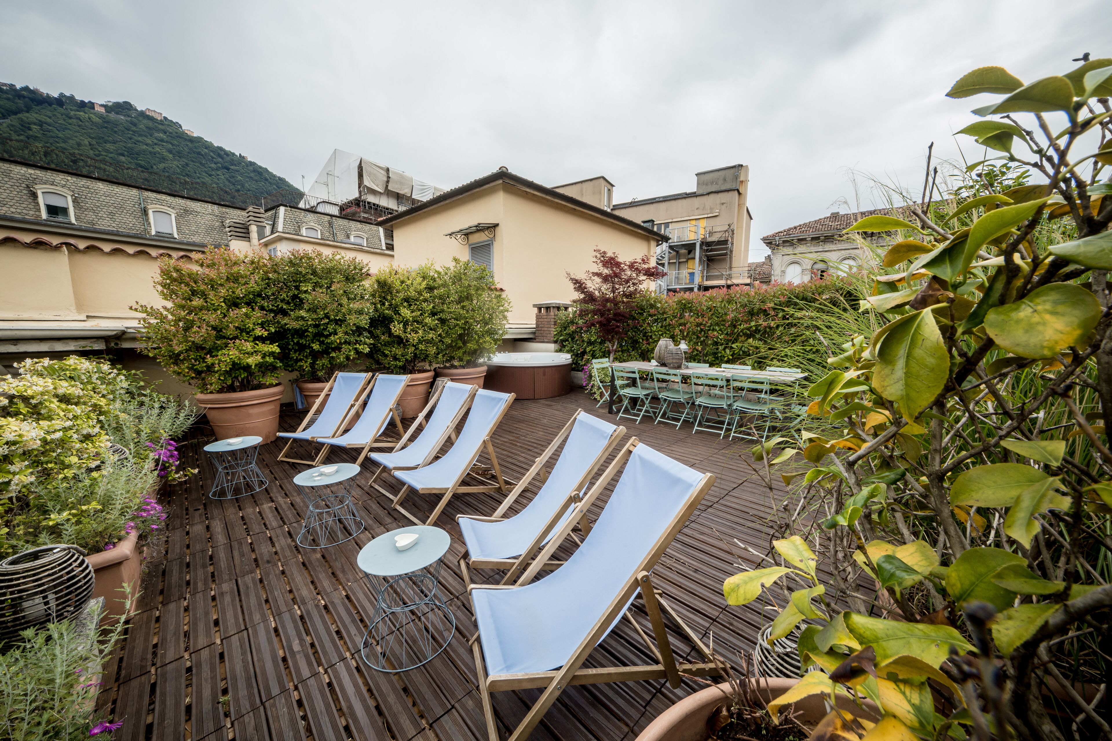 A rooftop terrace with lounge chairs and tables, surrounded by potted plants and a view of buildings.