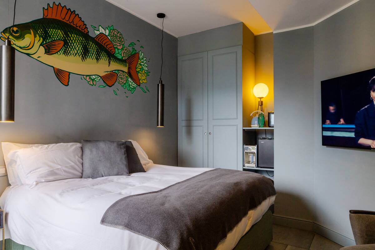 A hotel room with a bed, a wall-mounted fish mural, a television, and a desk with a lamp.
