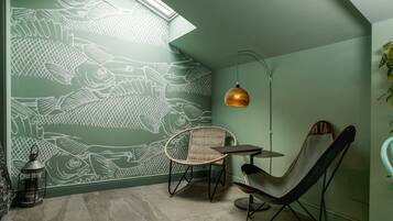 Una habitación con una pared verde decorada con un motivo de peces, una silla de mimbre, un sillón de cuero, una mesita y una lámpara colgante.