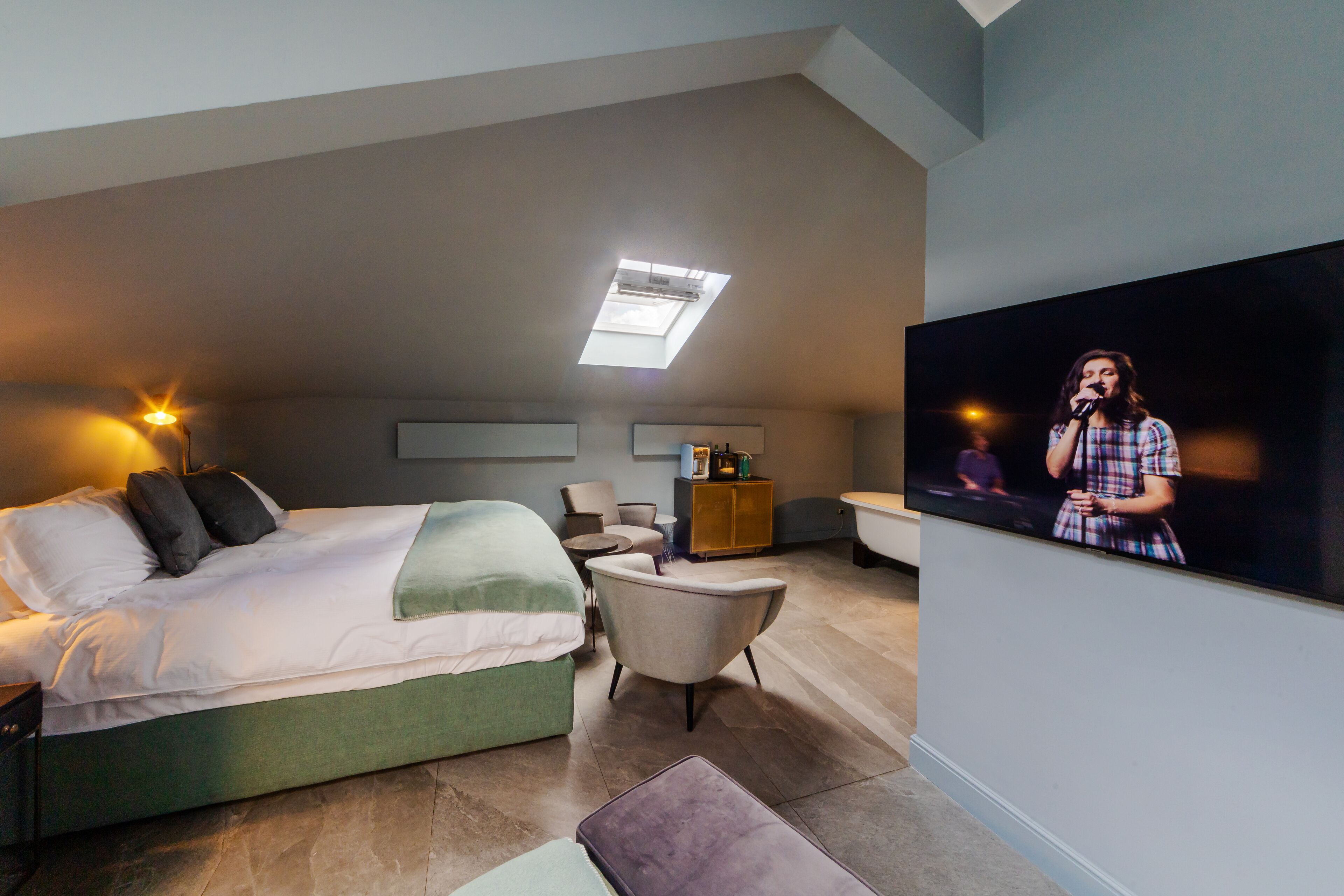 A modern hotel room with a bed, a seating area, a television, and a skylight.