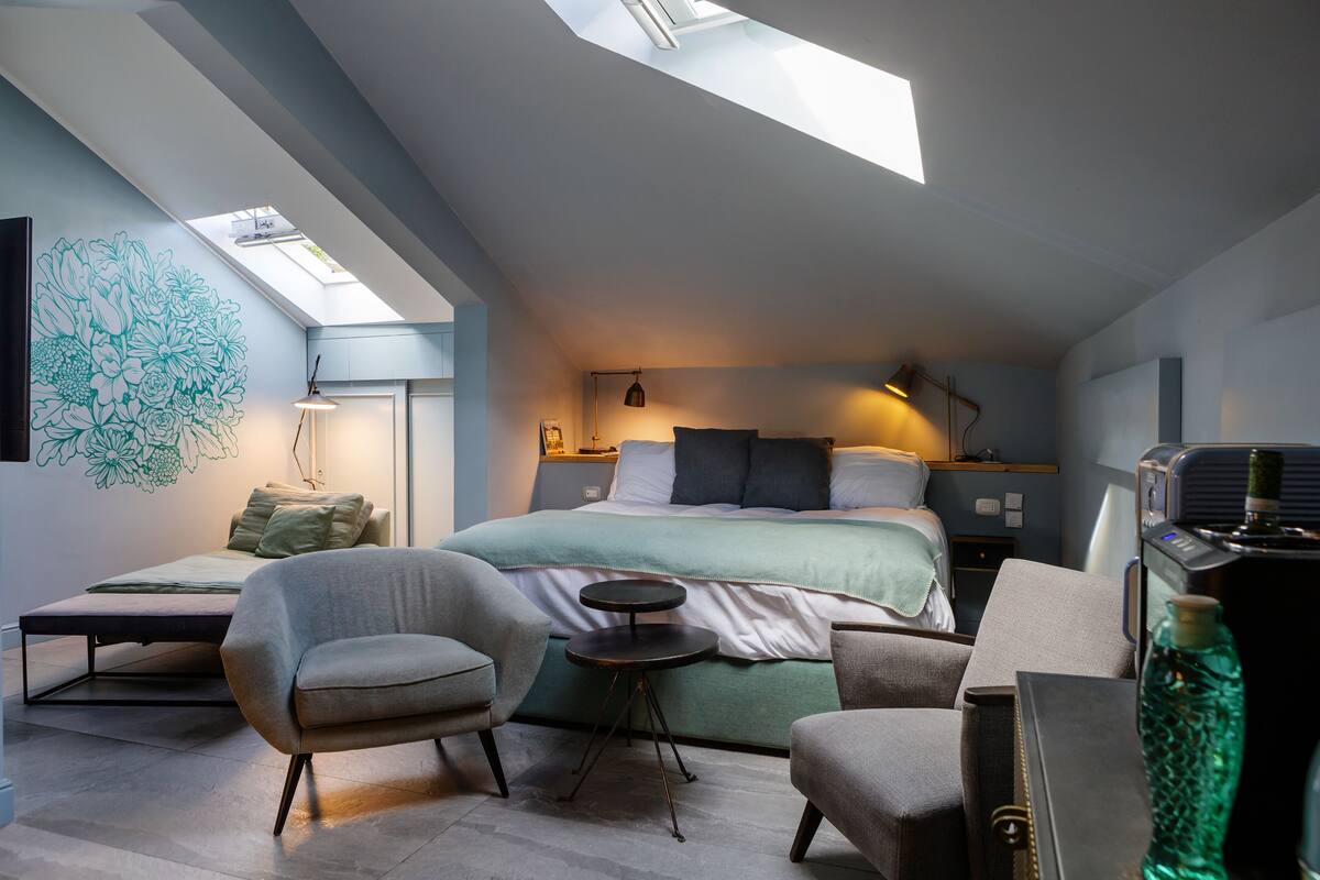 A modern bedroom with a skylight, a bed with a green bedspread, two armchairs, a small side table, and a nightstand with a lamp.
