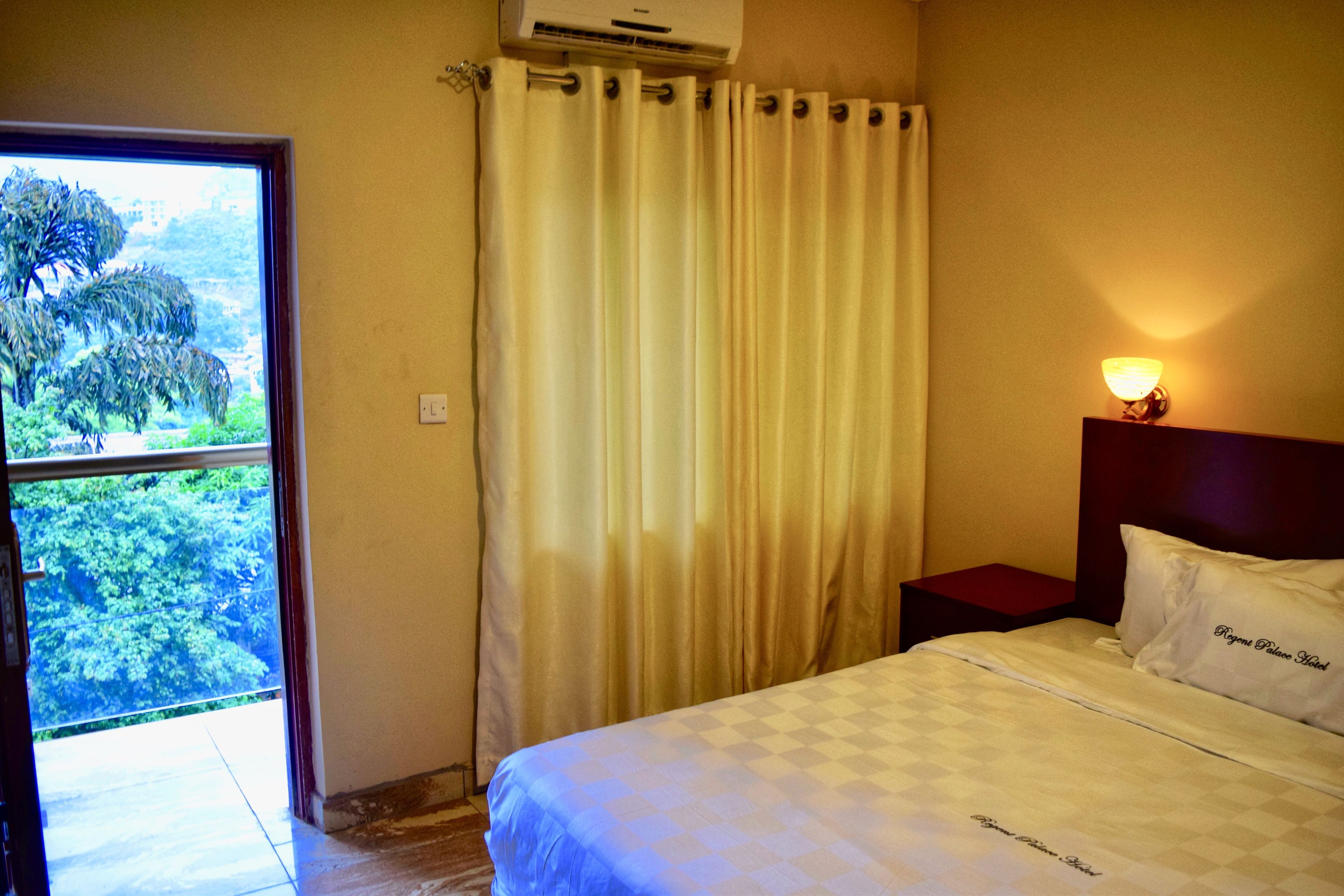 Habitación de hotel con cama, mesita de noche, lámpara y vistas a zonas verdes por una ventana.