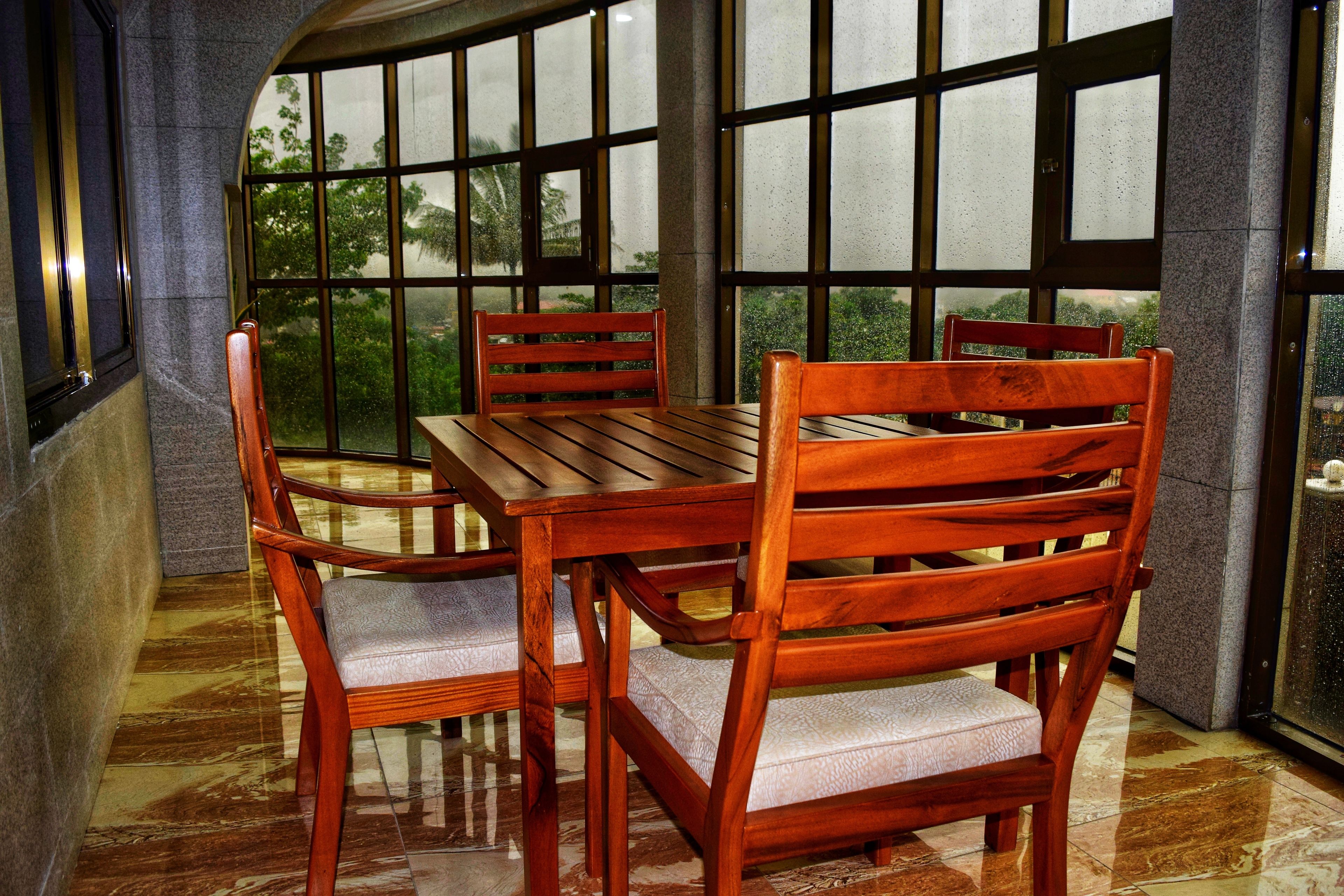 Una mesa y sillas de madera en una habitación con ventanas grandes y piso de baldosas.