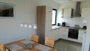 Cozinha moderna com armários brancos, fogão e máquina de café. Há uma mesa de jantar com cadeiras e uma janela acima.