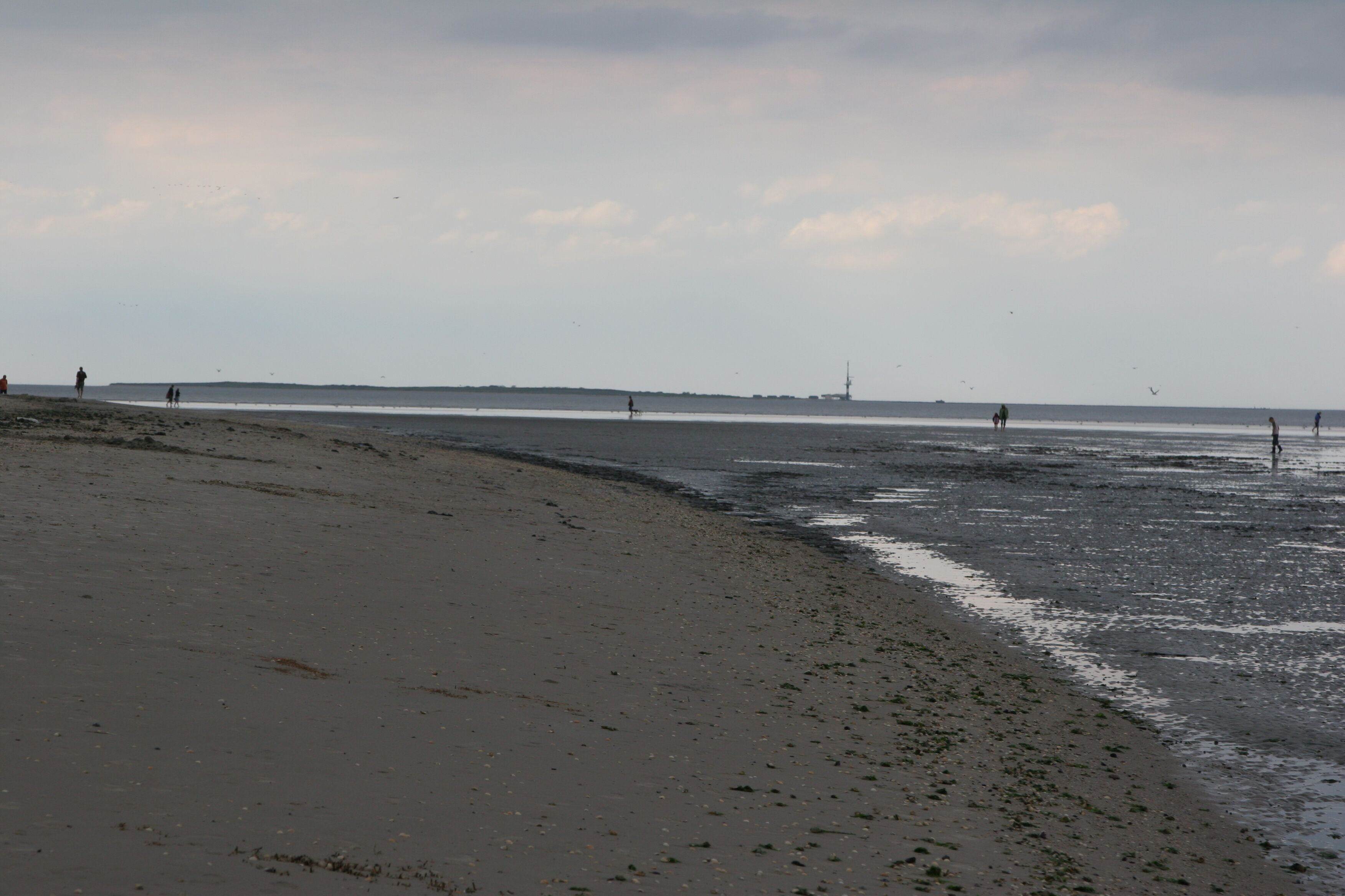 A beach with a wide sandy shore, a calm sea, and a distant shoreline with a few people and a structure.