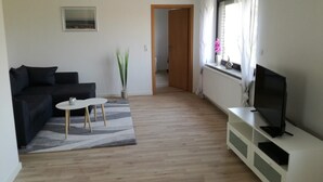 Uma sala de estar moderna com um sofá preto, uma mesinha branca, uma planta em vaso e uma televisão em um armário branco.