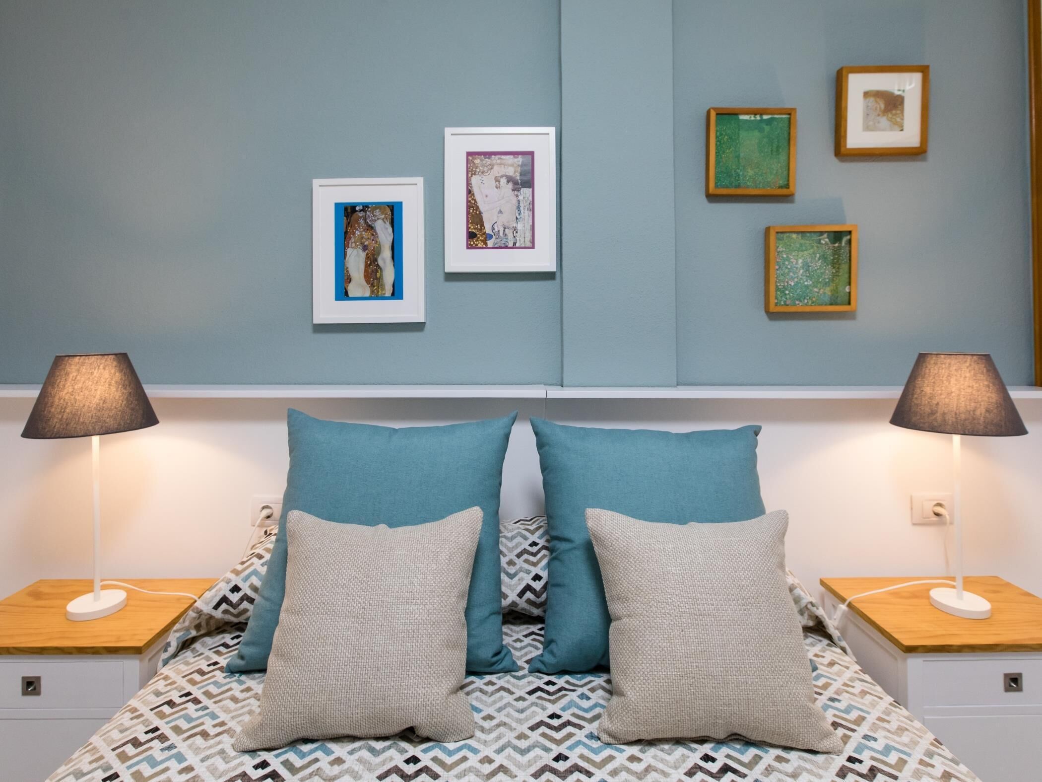 A bedroom with a bed, two bedside lamps, and four framed artworks on the wall.