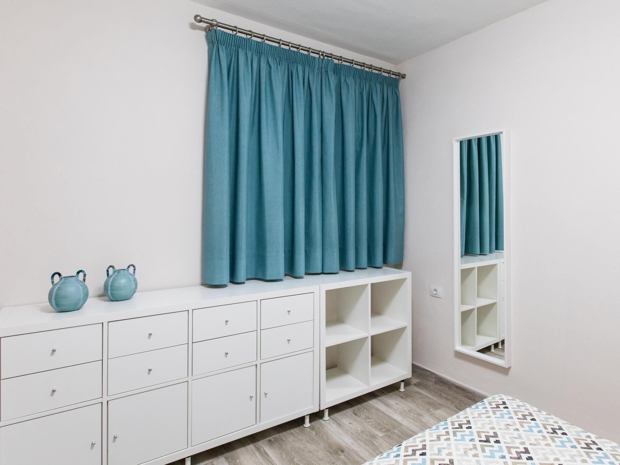 A bedroom with a white dresser, two blue vases, and a teal curtain.