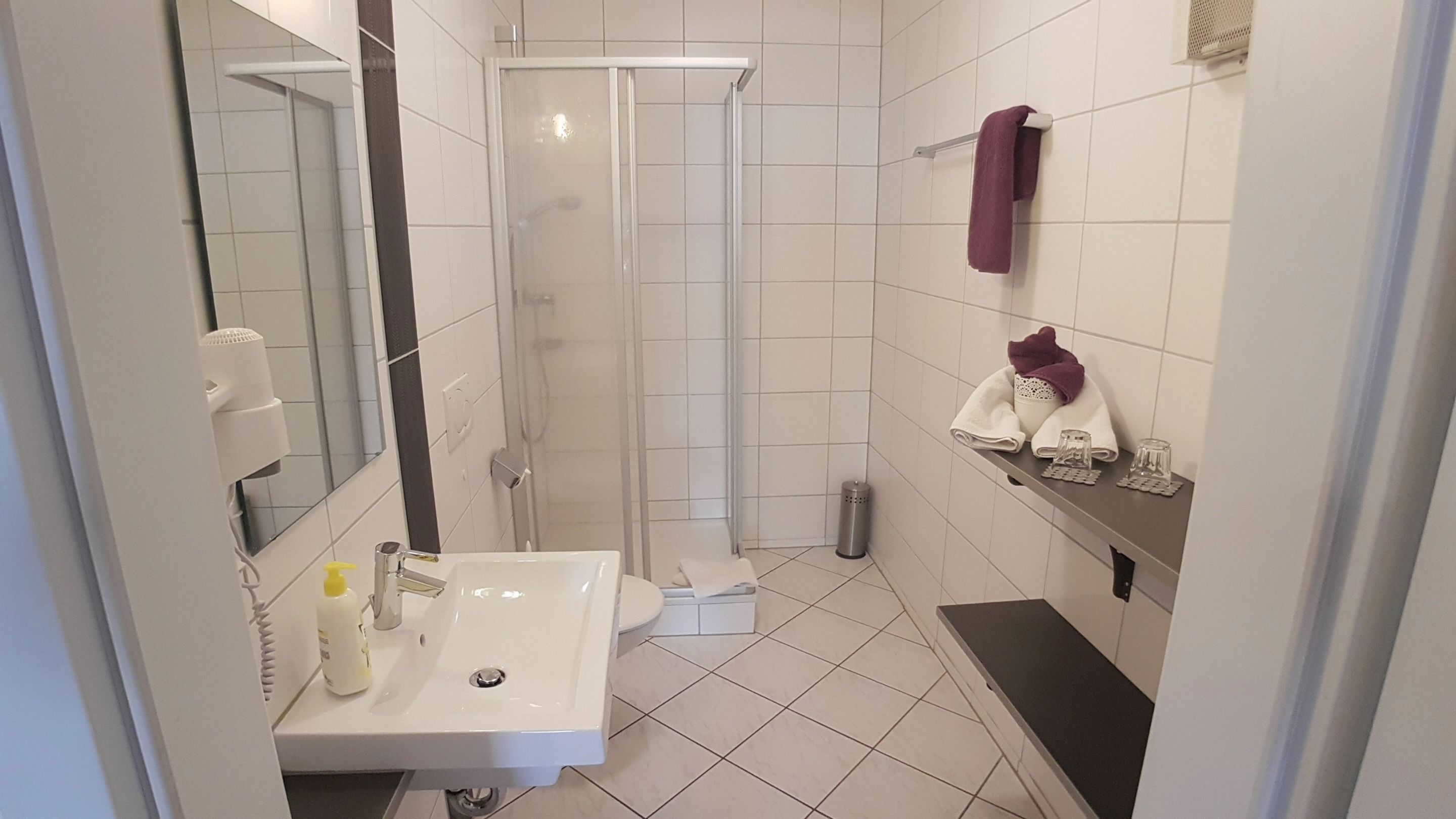 A modern bathroom with a white sink, mirror, and shower. There are towels and toiletries on a shelf.
