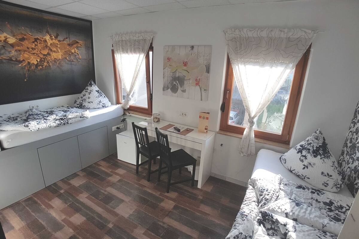 A compact room with a bed, a desk, and a chair. There are two windows with curtains, a painting on the wall, and a small cabinet.
