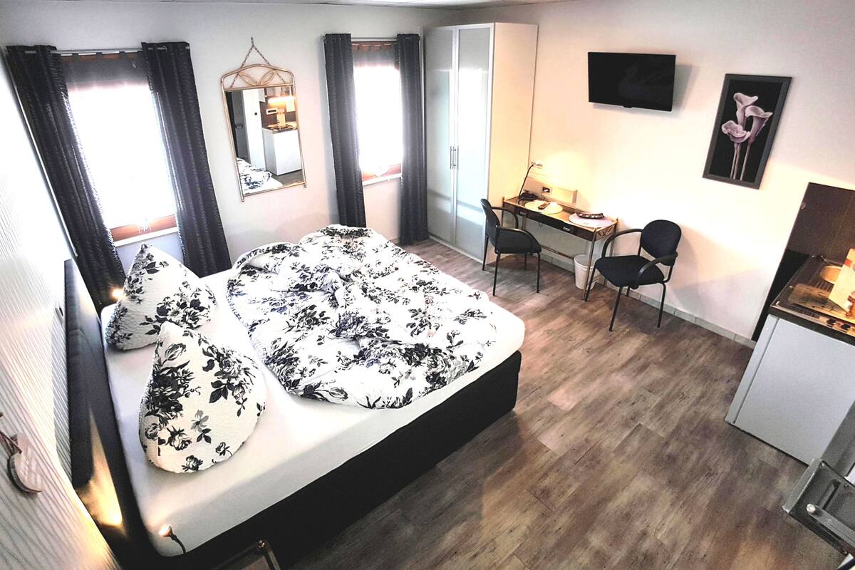A bedroom with a bed, a desk, two chairs, a mirror, and a TV on the wall.