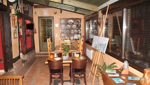 Un comedor con una mesa y sillas de madera, un caballete con un cuadro, y una repisa con vajilla.