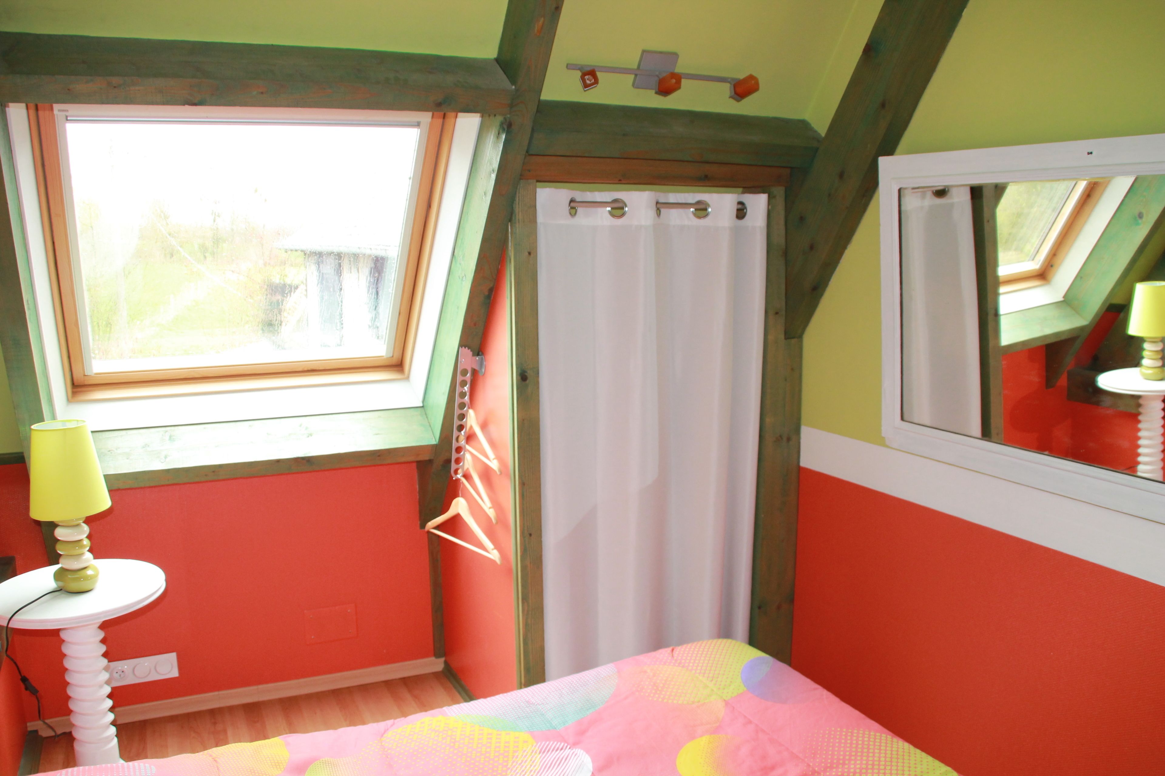 A bedroom with a skylight, a bed with a colorful bedspread, a bedside table with a lamp, and a window with a curtain.